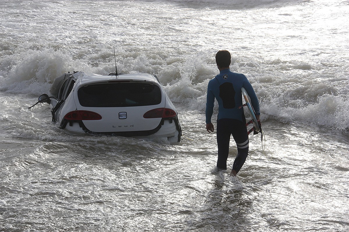 Un surfista, al costat d'un cotxe arrossegat per la pluja, aquest dijous al matí