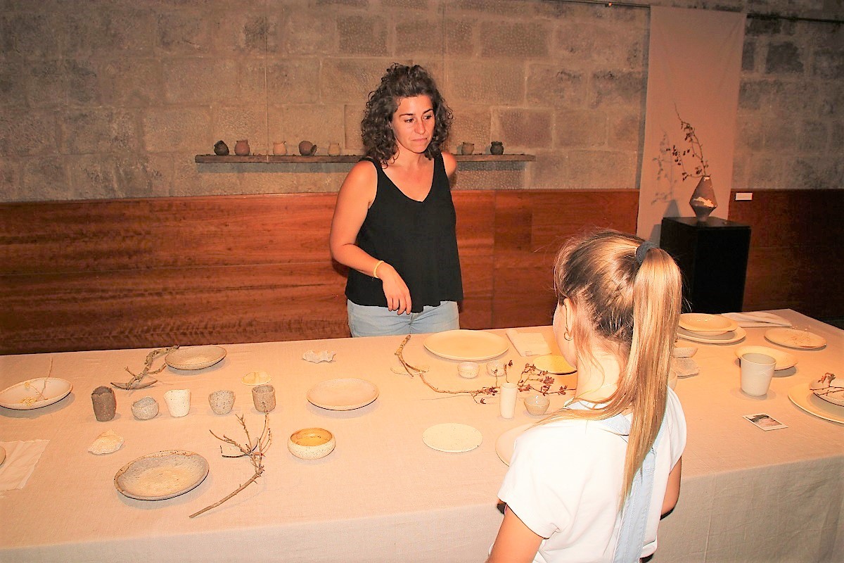 La Sara mostrant als visitants  una taula amb diferents peces de vaixella i ceràmica