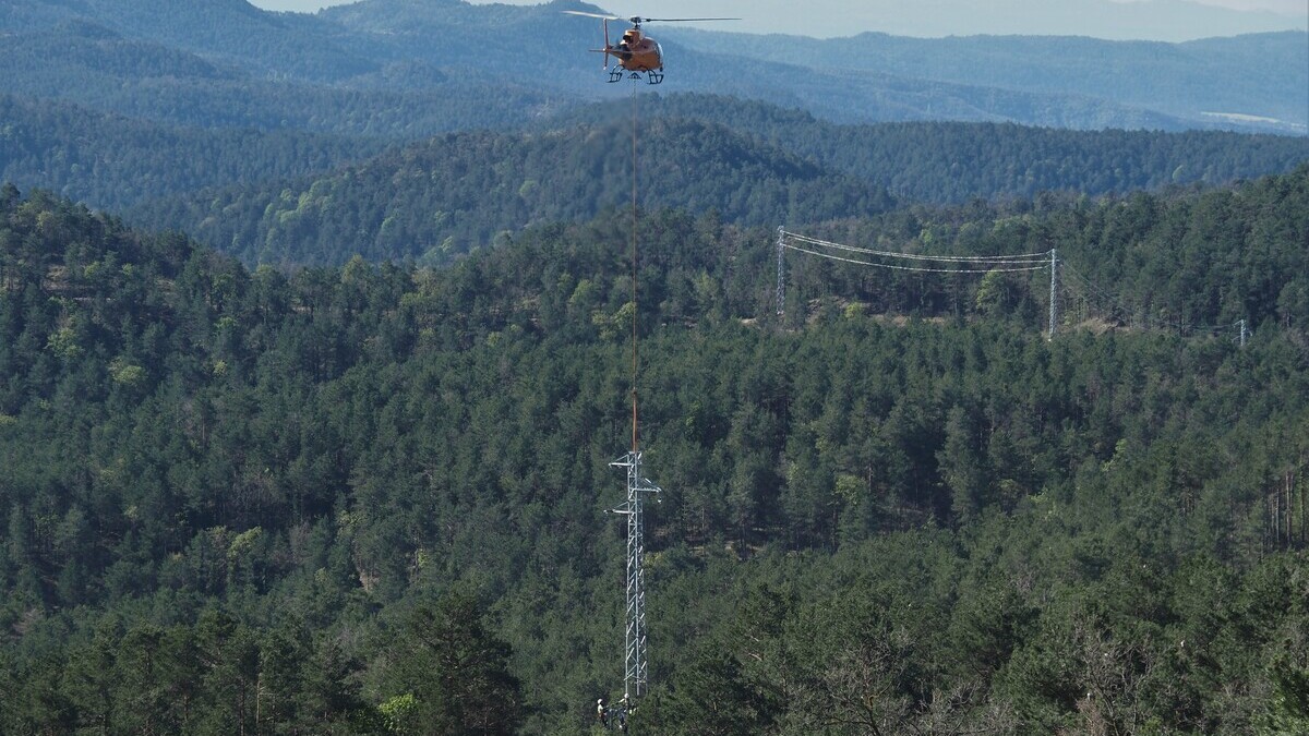 Un helicòpter i diversos operaris a peu treballen en la instal·lació d'una de les noves torres elèctriques